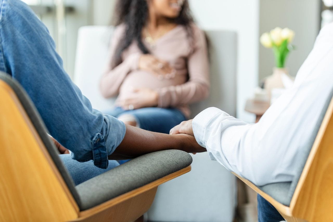 Couple holds hands for support in clinic waiting room, pregnant woman smiling with hand on belly in background.
