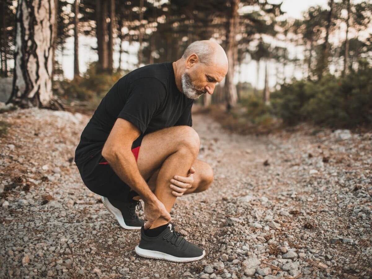 Older man outside, holds his right ankle as if in pain